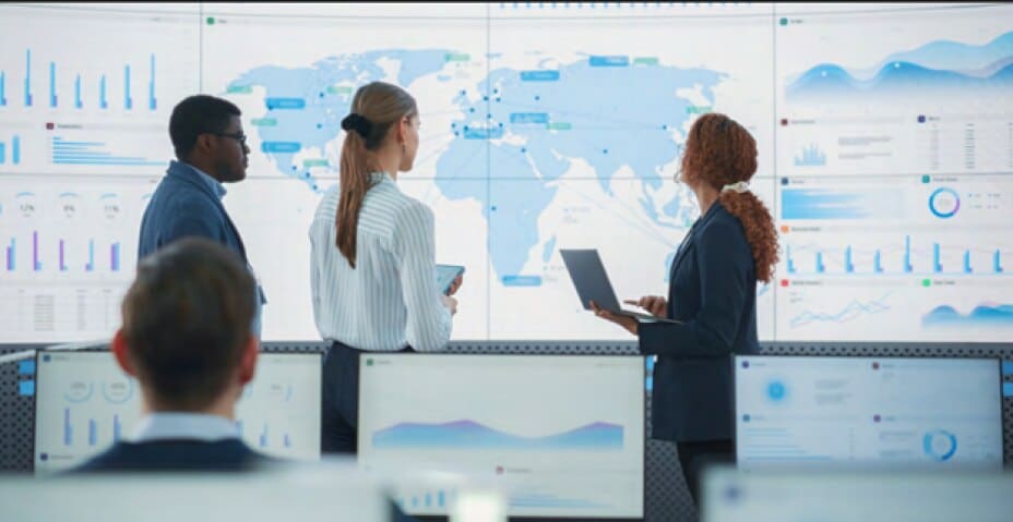 Three supply chain executives analyze supply chain data on a large map with several charts of data